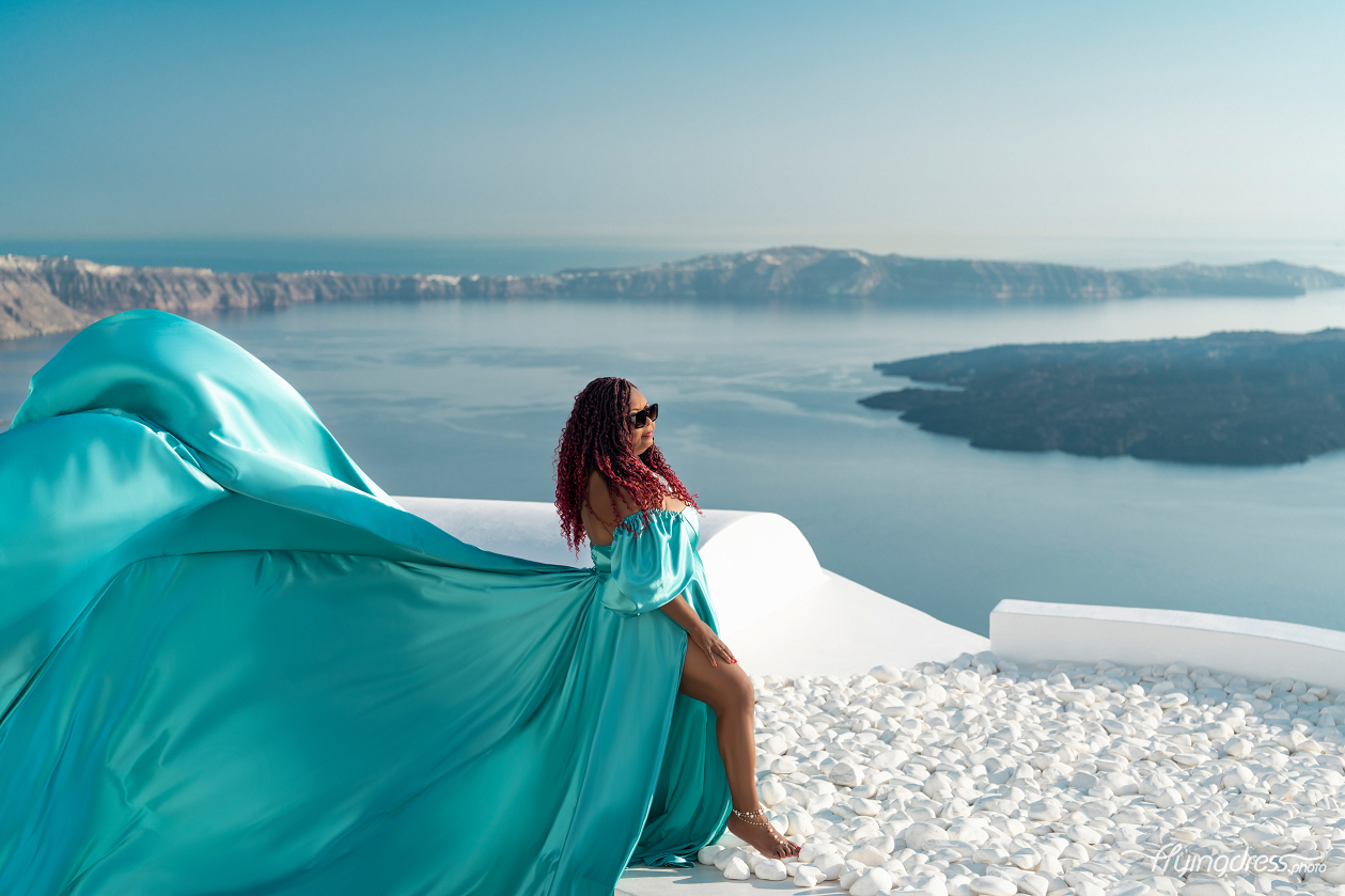 A woman in an emerald green dress sits elegantly on white pebbles, with the dress flowing behind her, overlooking a breathtaking view of calm waters and distant islands under a clear sky.